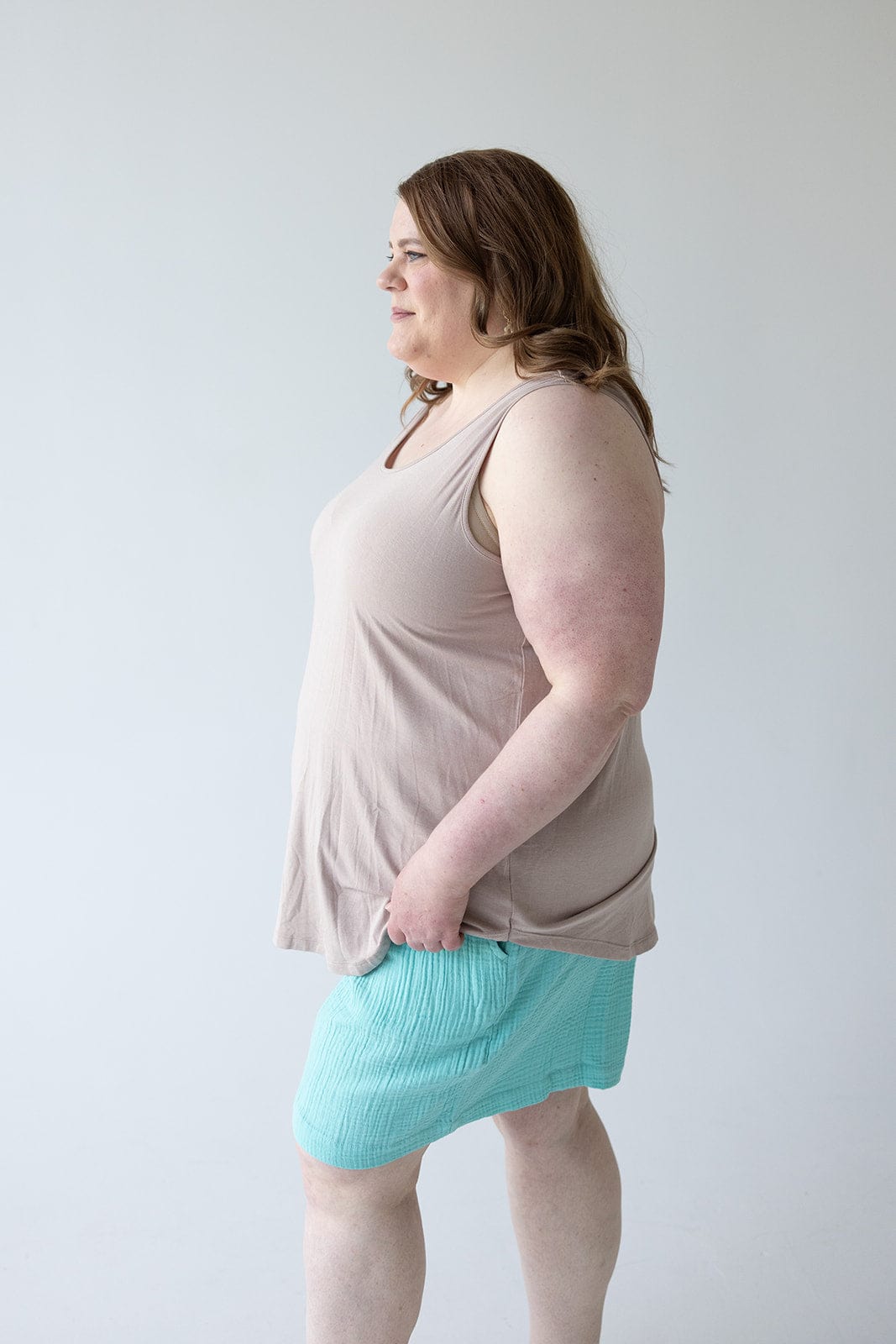 A person with long hair, wearing a chic and comfy BASIC ROUND NECK TUNIC TANK IN FROSTED TOFFEE by Love Marlow and turquoise shorts, stands against a plain white background, looking to the side with a hand in their pocket.