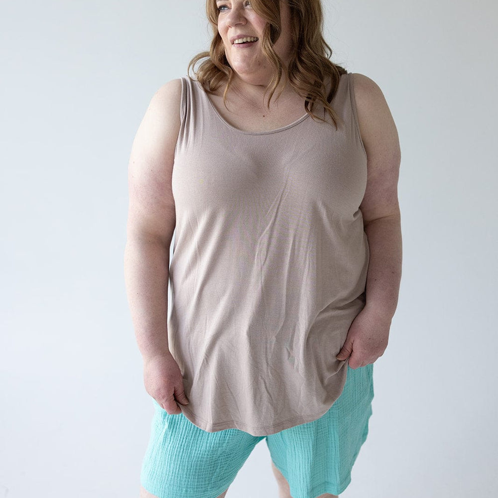 A woman with shoulder-length hair is standing and smiling in front of a plain white background, wearing a chic and comfy BASIC ROUND NECK TUNIC TANK IN FROSTED TOFFEE by Love Marlow paired with turquoise shorts.