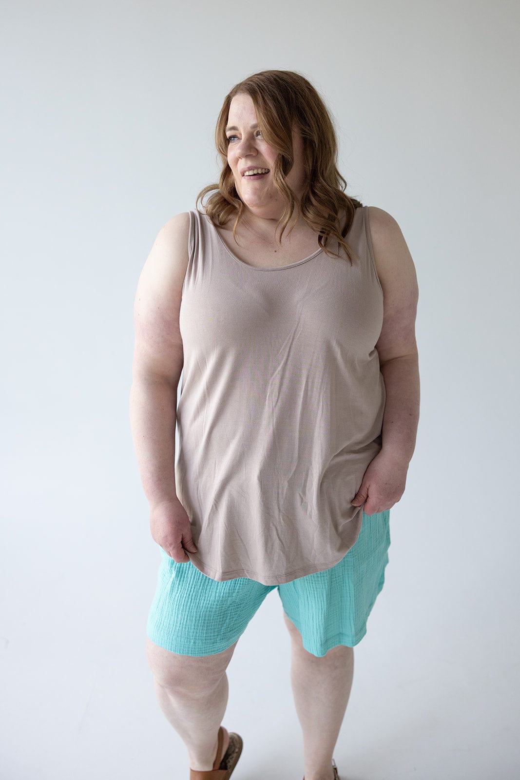 A woman with shoulder-length hair is standing and smiling in front of a plain white background, wearing a chic and comfy BASIC ROUND NECK TUNIC TANK IN FROSTED TOFFEE by Love Marlow paired with turquoise shorts.