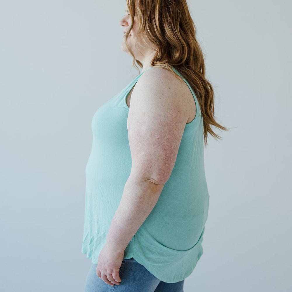 A person with long hair wearing a light green, BASIC ROUND NECK TUNIC TANK IN REEF from Love Marlow made of ultra-soft Modal fabric and blue jeans stands sideways against a neutral background.