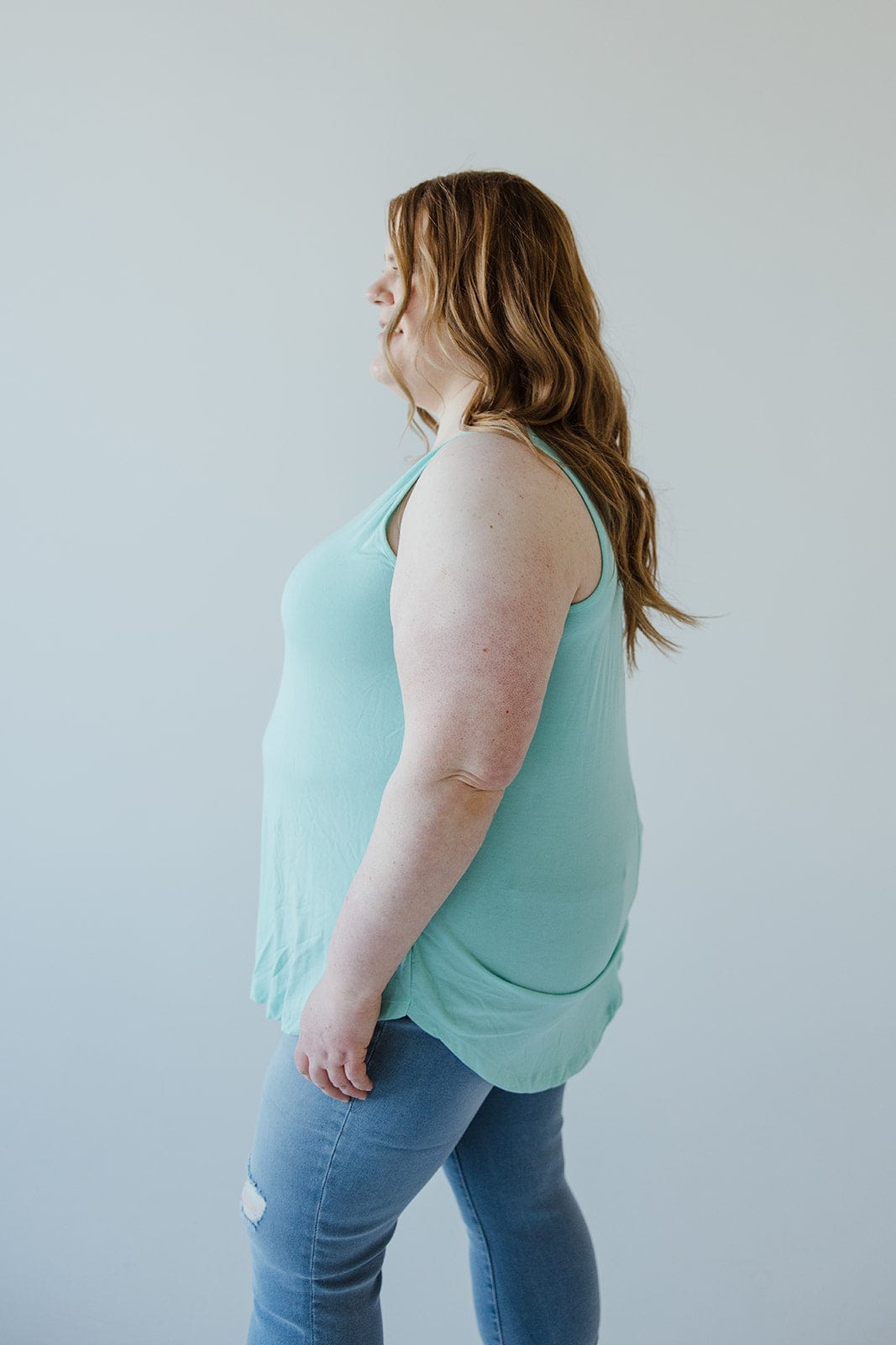 A person with long hair wearing a light green, BASIC ROUND NECK TUNIC TANK IN REEF from Love Marlow made of ultra-soft Modal fabric and blue jeans stands sideways against a neutral background.