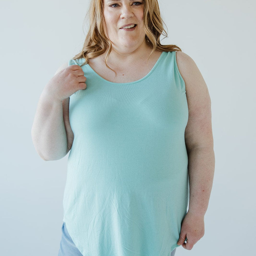 A woman stands against a plain background, wearing a light blue BASIC ROUND NECK TUNIC TANK IN REEF by Love Marlow with a high-low rounded hem and ripped jeans. She has her right hand up near her shoulder.