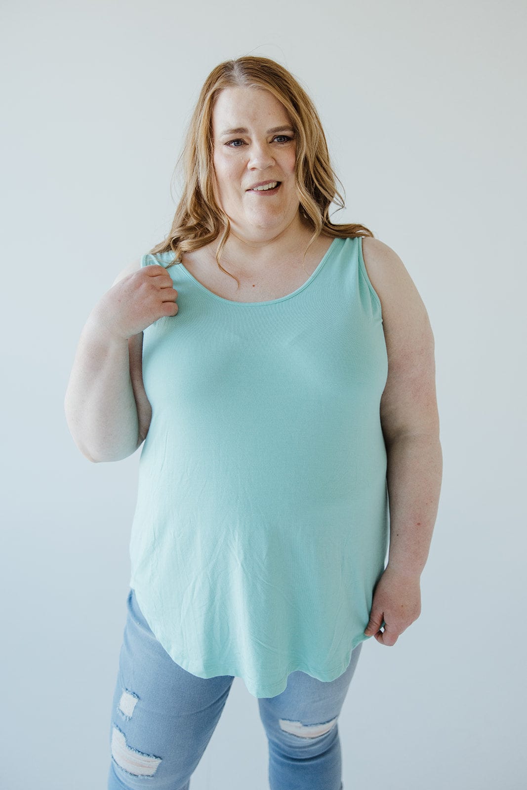 A woman stands against a plain background, wearing a light blue BASIC ROUND NECK TUNIC TANK IN REEF by Love Marlow with a high-low rounded hem and ripped jeans. She has her right hand up near her shoulder.