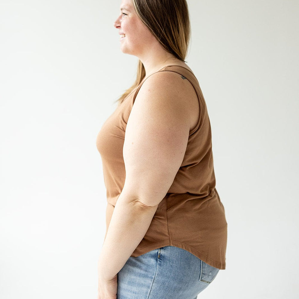 A person with long hair wearing a Love Marlow BASIC ROUND NECK TUNIC TANK IN URBAN BRONZE and blue jeans, made of soft modal fabric, stands sideways against a white background.