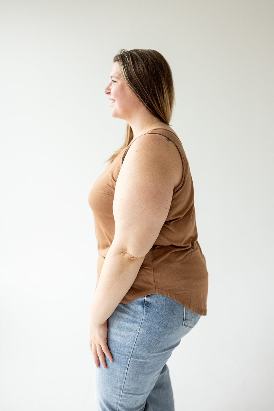 A person with long hair wearing a Love Marlow BASIC ROUND NECK TUNIC TANK IN URBAN BRONZE and blue jeans, made of soft modal fabric, stands sideways against a white background.