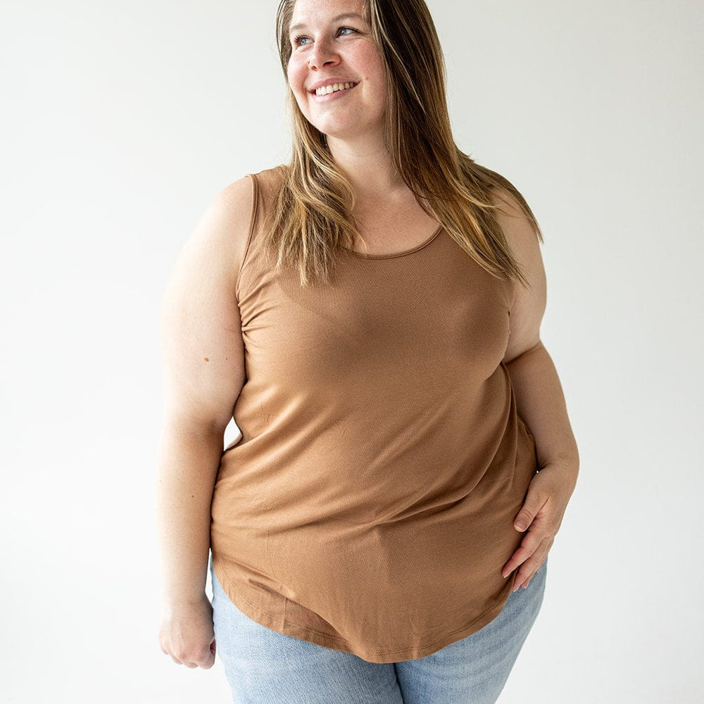 A person with long brown hair, wearing a BASIC ROUND NECK TUNIC TANK IN URBAN BRONZE from Love Marlow and light blue jeans, stands smiling against a plain white background.