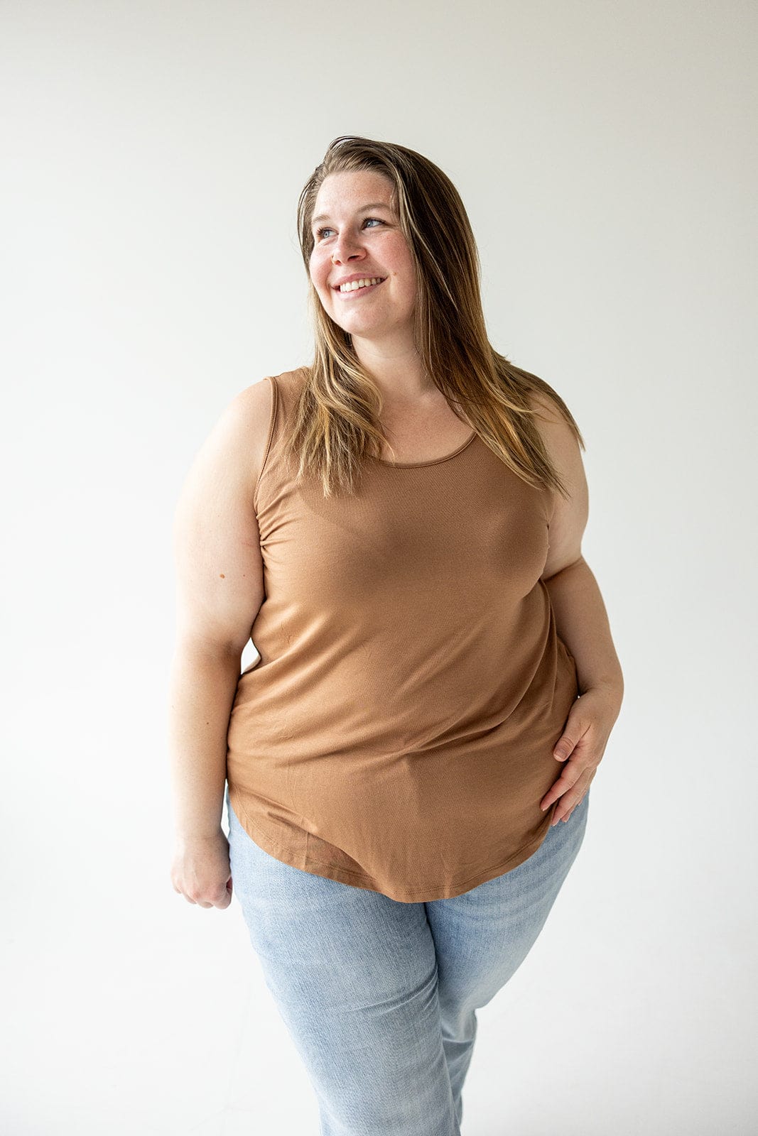 A person with long brown hair, wearing a BASIC ROUND NECK TUNIC TANK IN URBAN BRONZE from Love Marlow and light blue jeans, stands smiling against a plain white background.