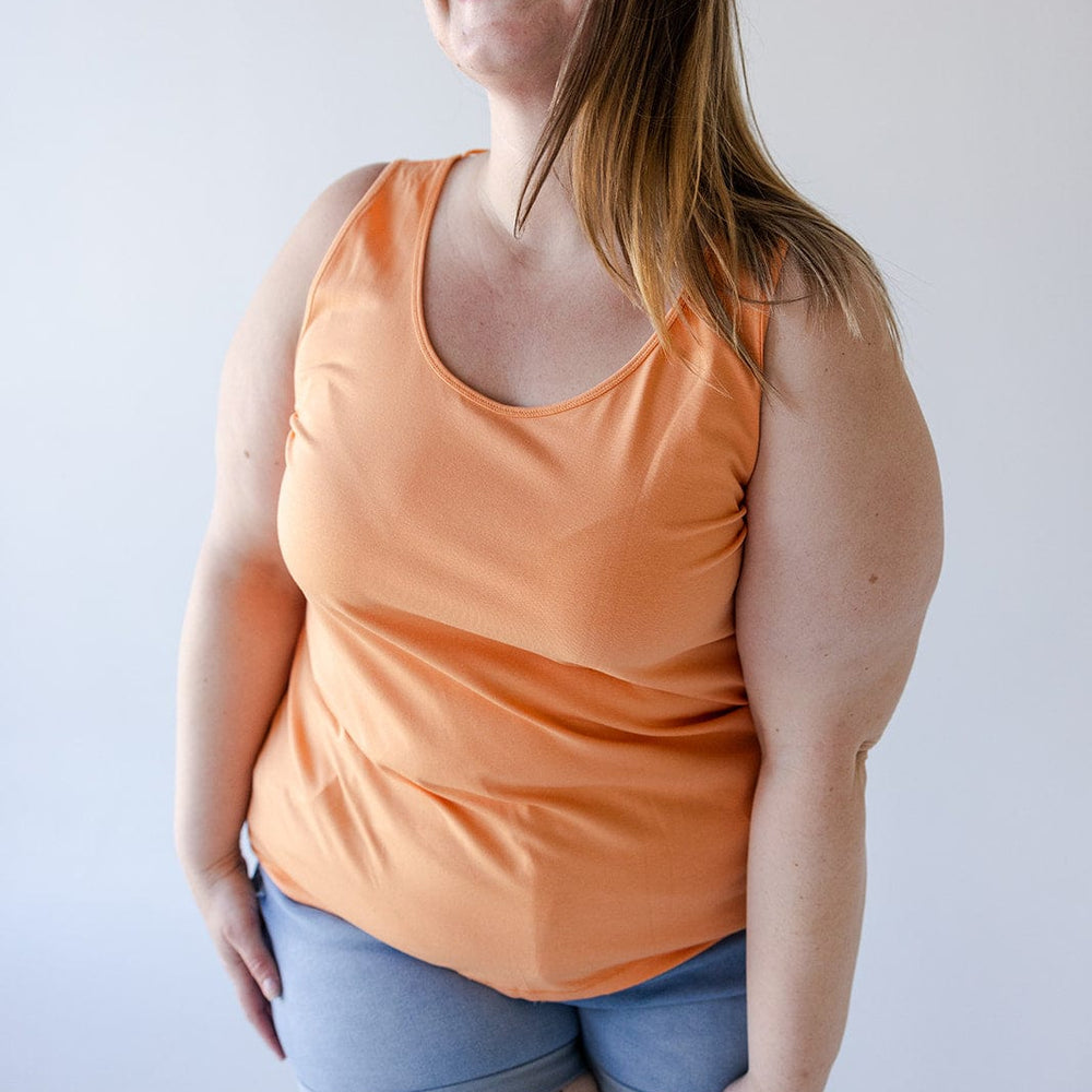 A person wearing glasses, a vibrant BASIC SCOOP NECK TANK IN TANGERINE by Love Marlow, and blue shorts smiles with their eyes closed. They have long hair and a floral tattoo on their thigh.