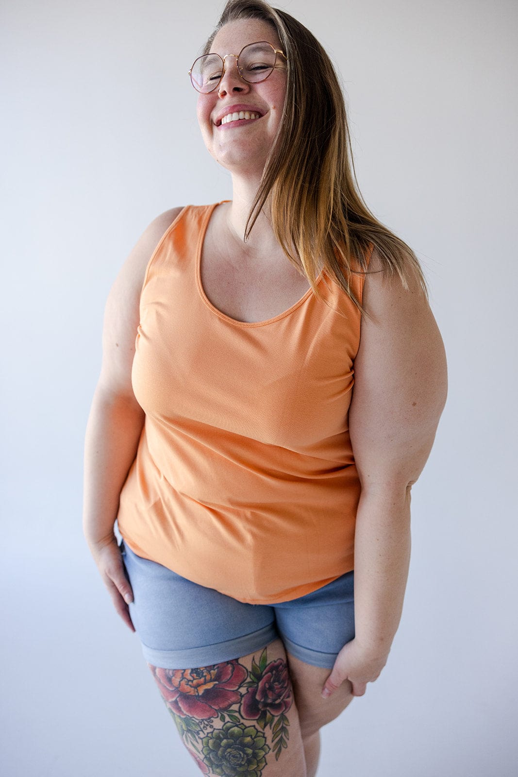 A person wearing glasses, a vibrant BASIC SCOOP NECK TANK IN TANGERINE by Love Marlow, and blue shorts smiles with their eyes closed. They have long hair and a floral tattoo on their thigh.