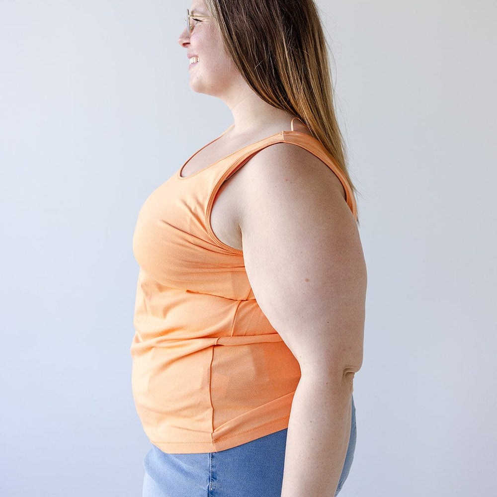 A woman with long hair and glasses is wearing a vibrant BASIC SCOOP NECK TANK IN TANGERINE by Love Marlow, made from a cotton-modal blend, paired with denim shorts. She stands in profile against a plain white background.