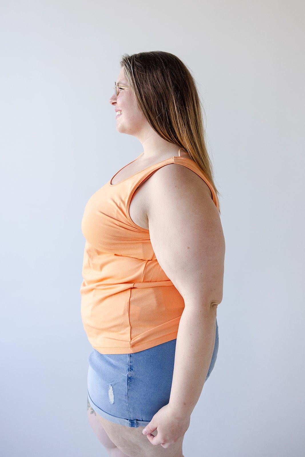 A woman with long hair and glasses is wearing a vibrant BASIC SCOOP NECK TANK IN TANGERINE by Love Marlow, made from a cotton-modal blend, paired with denim shorts. She stands in profile against a plain white background.