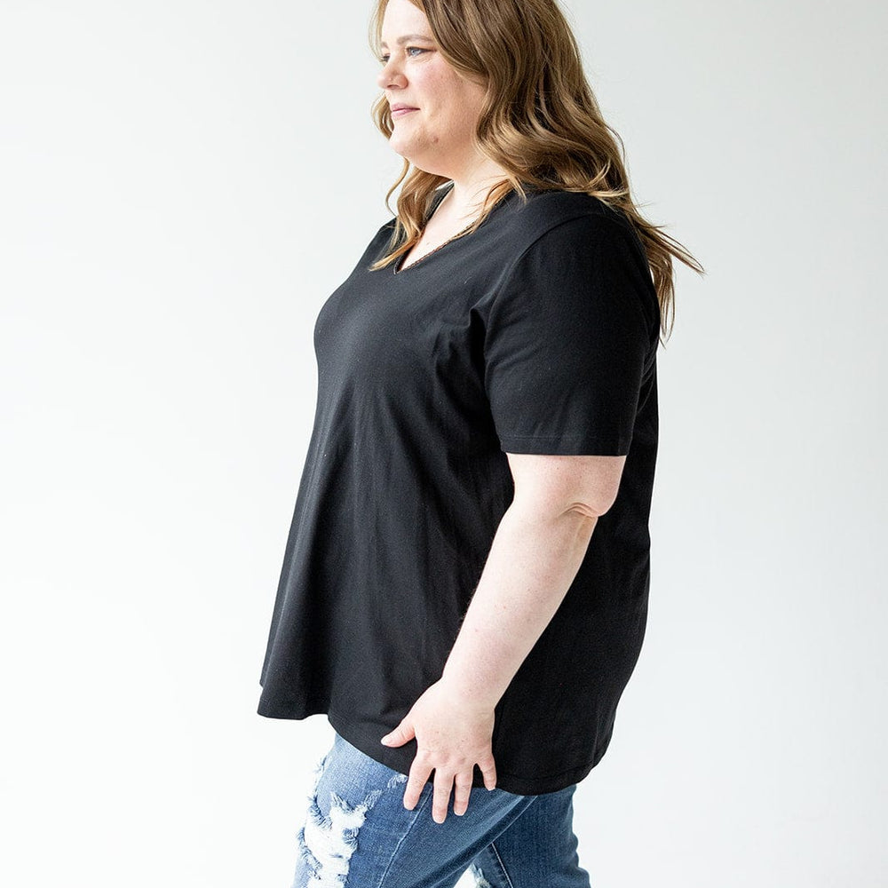 A woman in a BLACK BASIC V-NECK TEE WITH BRONZE SPARKLE by Love Marlow and ripped jeans stands against a light background, looking to her right.