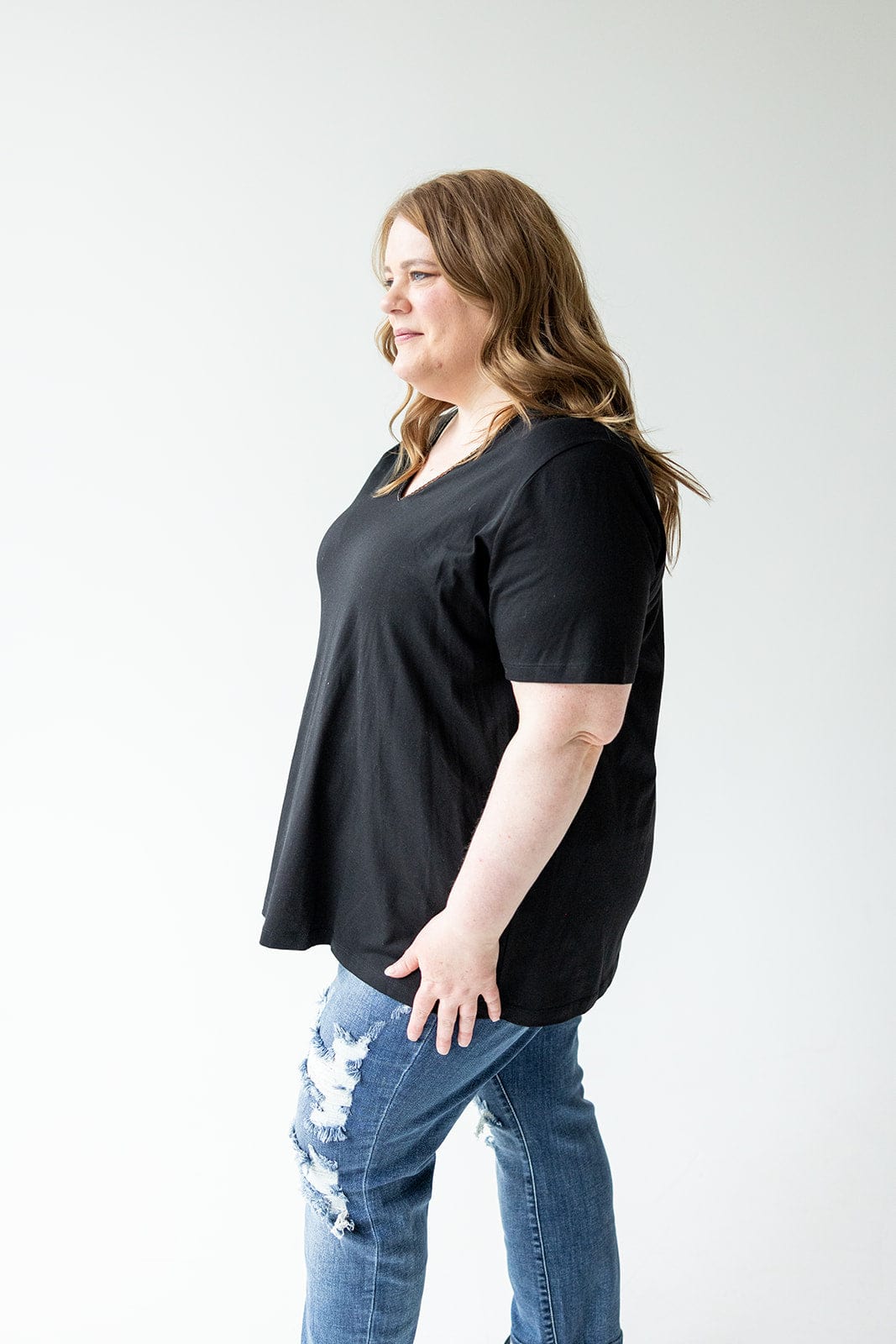 A woman in a BLACK BASIC V-NECK TEE WITH BRONZE SPARKLE by Love Marlow and ripped jeans stands against a light background, looking to her right.