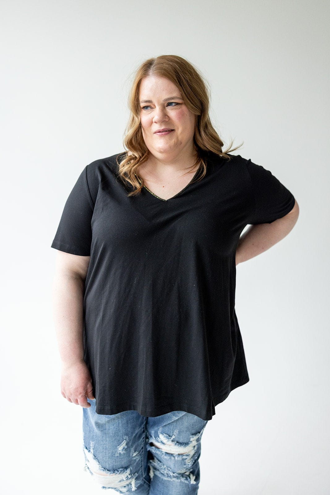 A person with long hair, wearing Love Marlow's BASIC V-NECK TEE WITH BRONZE SPARKLE IN BLACK and ripped jeans, stands against a plain white background.