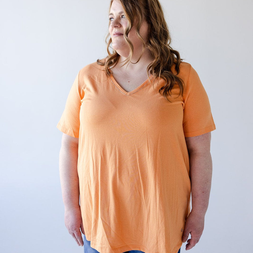 Person with long wavy hair wearing a BASIC V-NECK TEE WITH SPARKLE IN TANGERINE by Love Marlow, denim shorts, and sunglasses on their head, standing against a plain background.