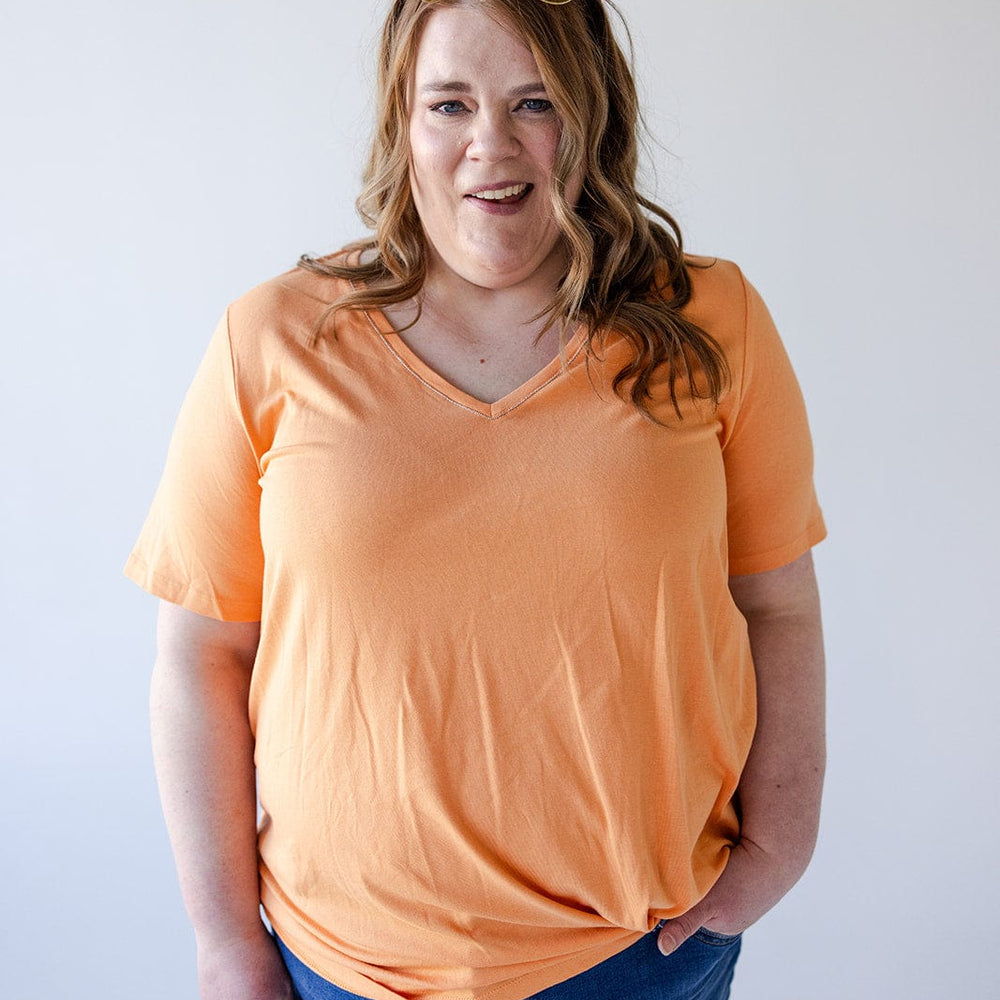 A woman with long hair wearing a BASIC V-NECK TEE WITH SPARKLE IN TANGERINE by Love Marlow, blue jean shorts, and sunglasses on her head stands against a plain white background.