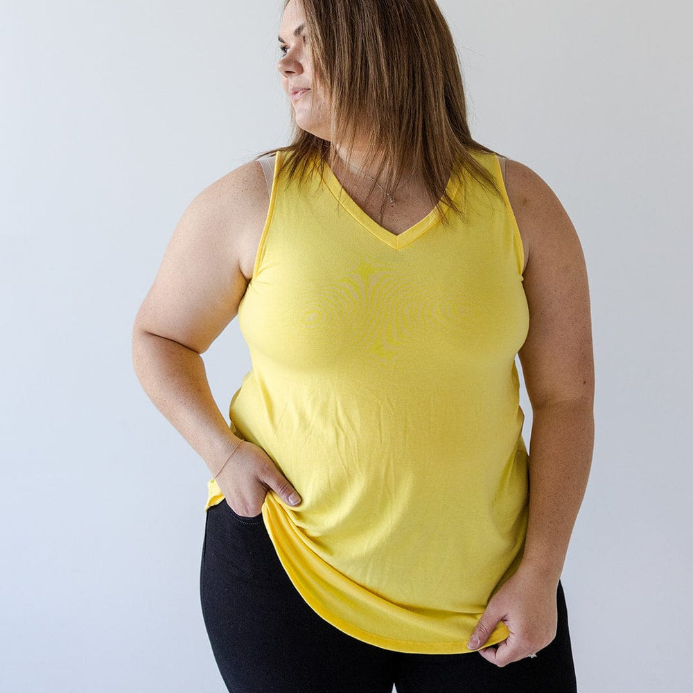 A person stands against a plain background, wearing the BASIC V-NECK TUNIC TANK IN SUNSHINE by Love Marlow and black pants, looking to the side with one hand holding the hem of their top.