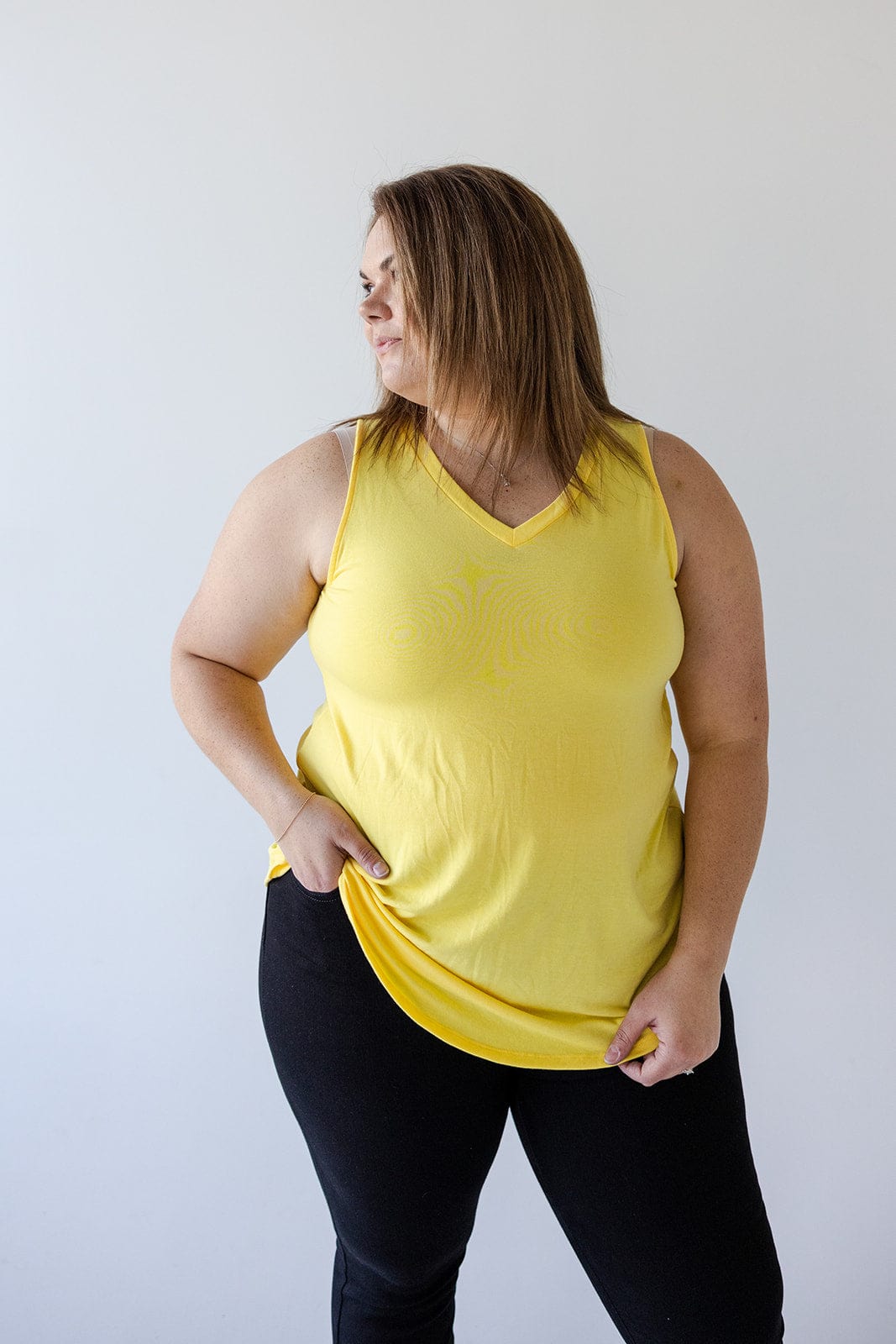 A person stands against a plain background, wearing the BASIC V-NECK TUNIC TANK IN SUNSHINE by Love Marlow and black pants, looking to the side with one hand holding the hem of their top.