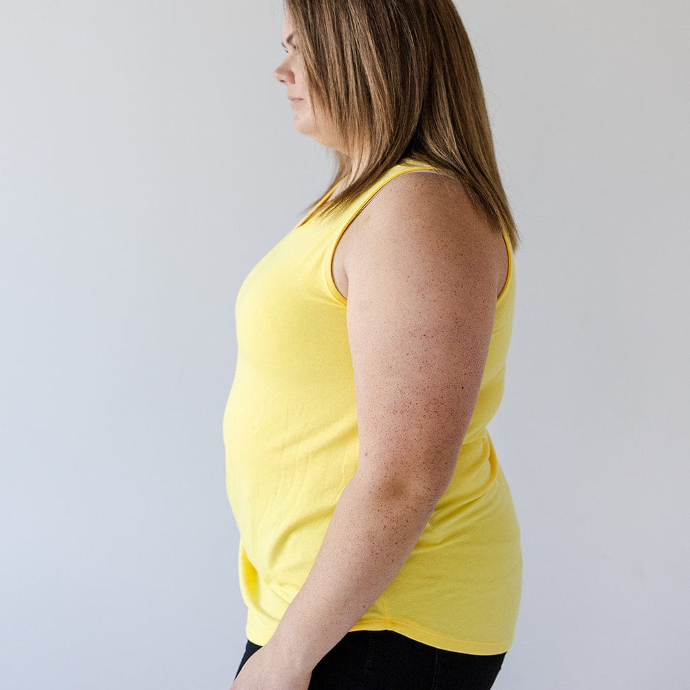 A person with medium-length hair, wearing the BASIC V-NECK TUNIC TANK IN SUNSHINE by Love Marlow made of super soft knit fabric and black pants, stands in profile against a plain background.