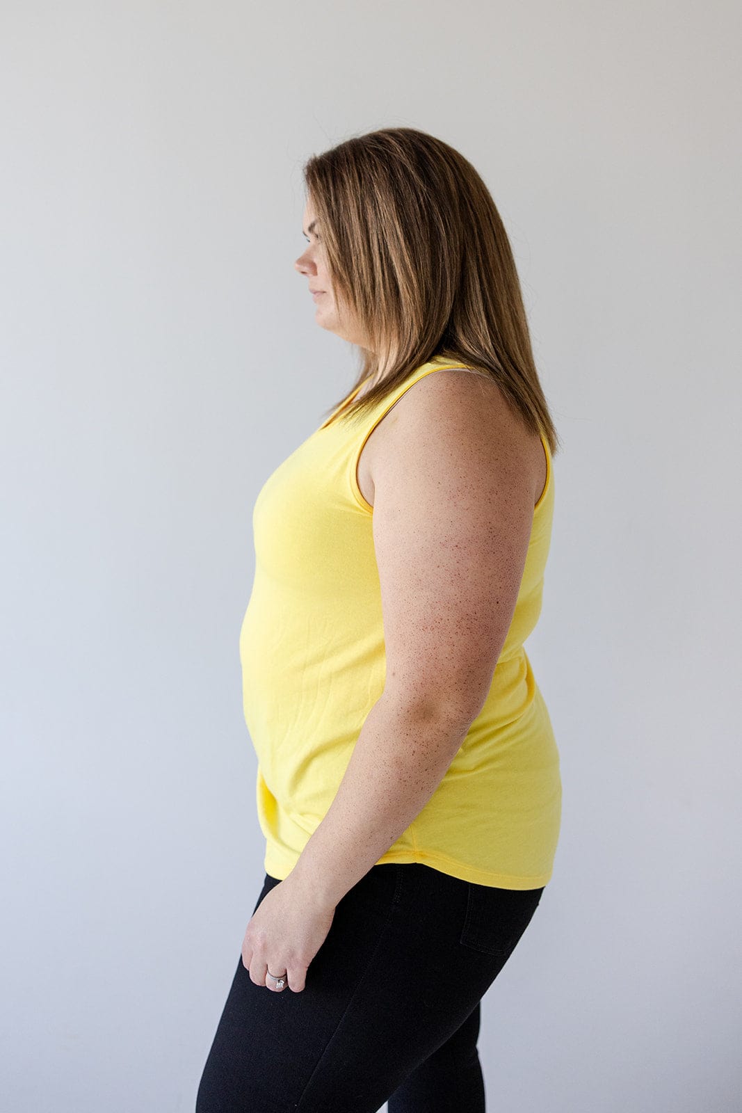 A person with medium-length hair, wearing the BASIC V-NECK TUNIC TANK IN SUNSHINE by Love Marlow made of super soft knit fabric and black pants, stands in profile against a plain background.