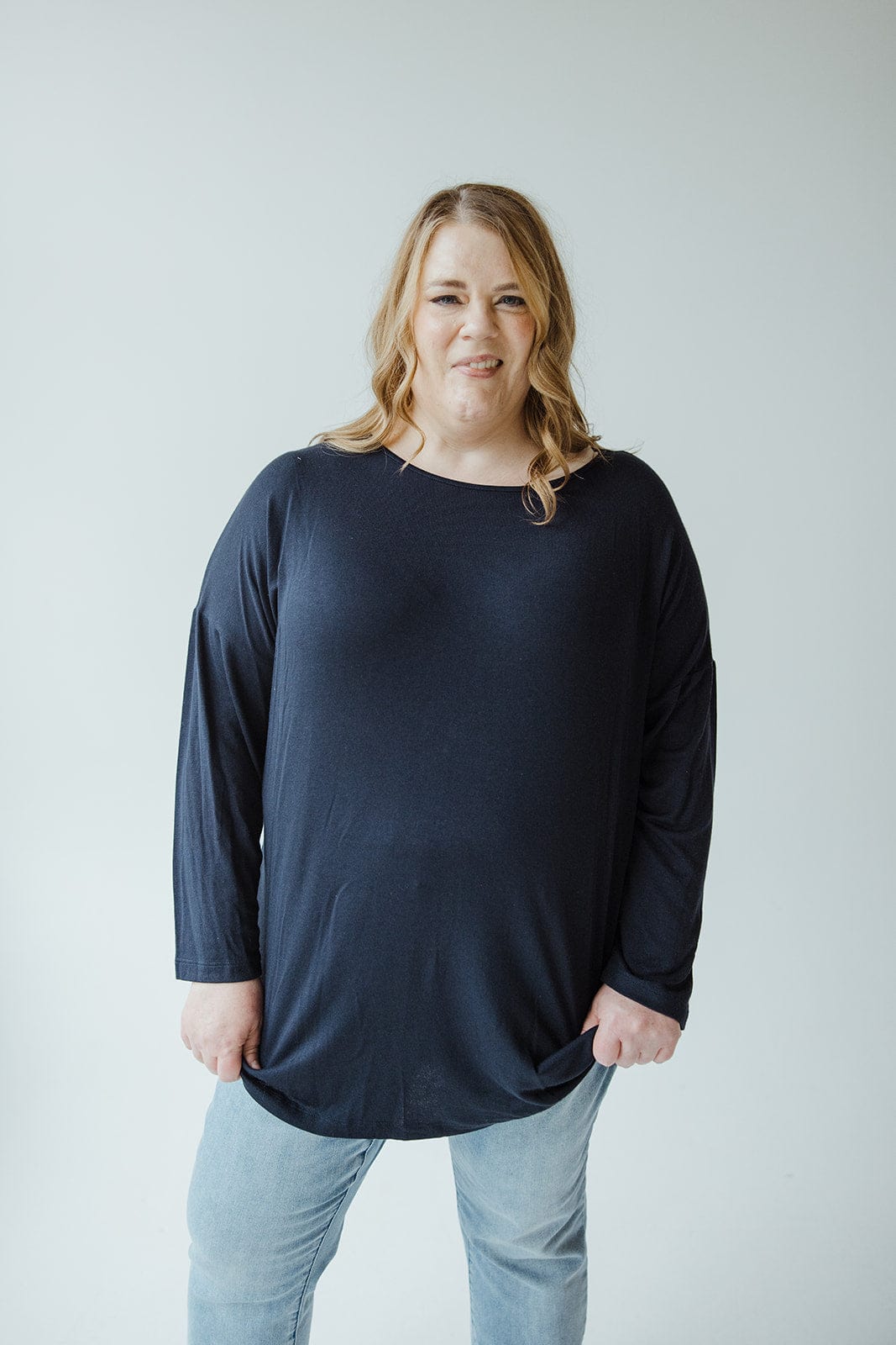 A person with long hair in a navy blue long-sleeved tee and blue jeans stands in front of a plain white background.