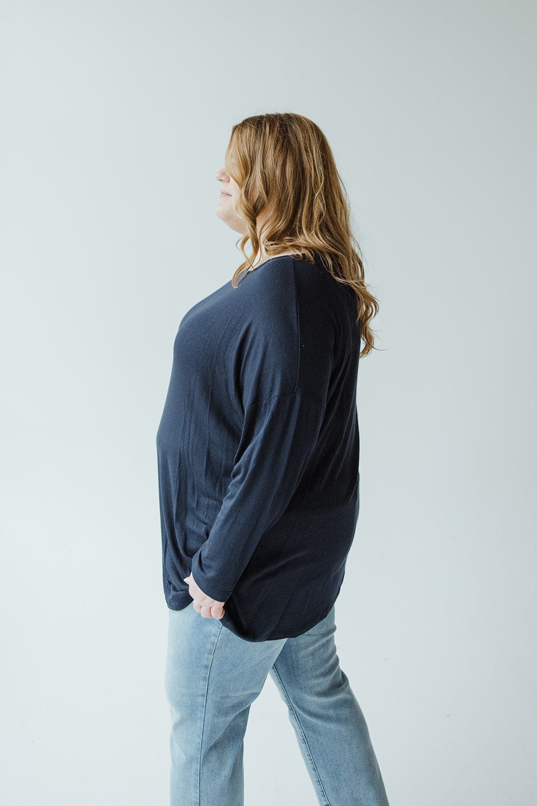 A person with long hair, dressed in a classic tunic made of lightweight fabric and jeans, stands sideways against a plain background.