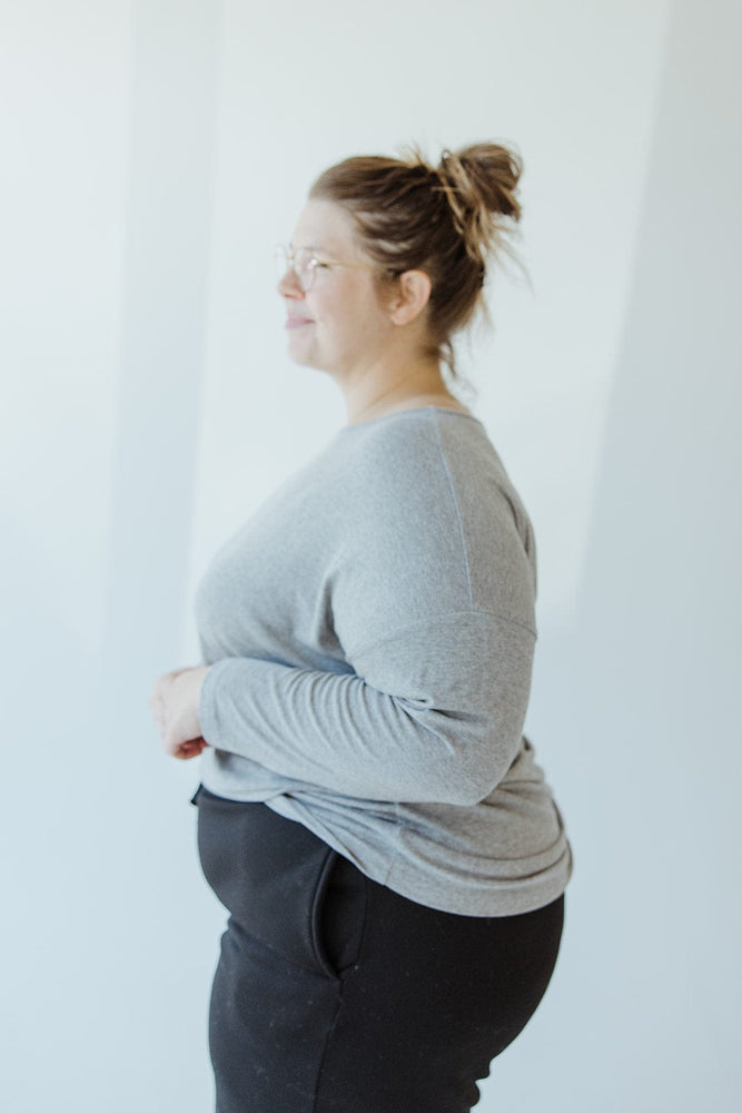 A person with their back to the camera, wearing black pants and a Yesta BASIC DOLMAN TUNIC in HEATHER GREY. Their hair is tied up in a messy bun.