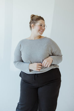 A person with glasses, wearing the BASIC DOLMAN TUNIC IN HEATHER GREY by Yesta and black pants made from a luxurious fabric, gazes to the side with a slight smile.