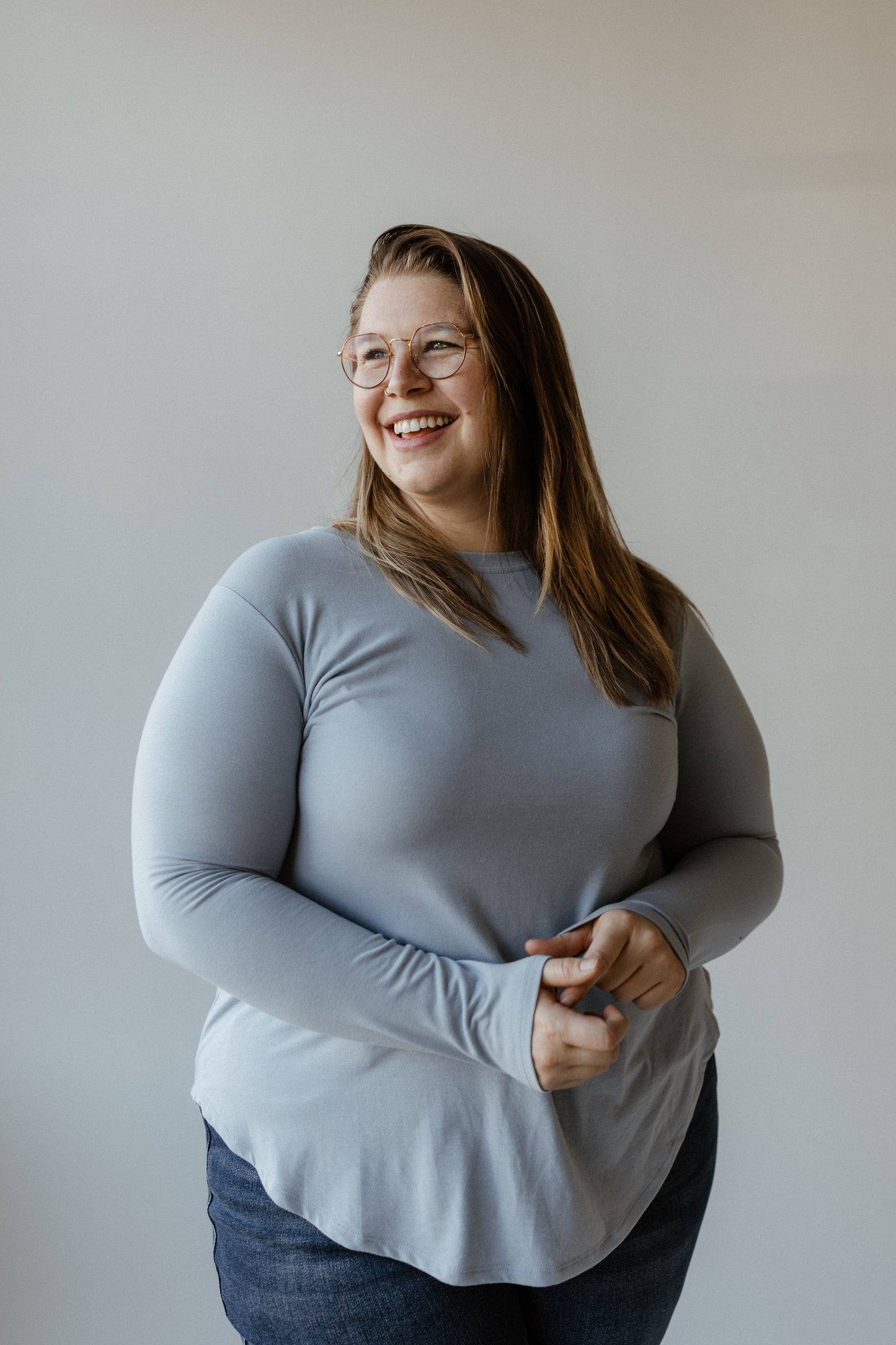 A person with long hair and glasses, wearing a light gray sweater made of super soft knit fabric, paired with dark jeans, is smiling while looking to the side against a plain background.