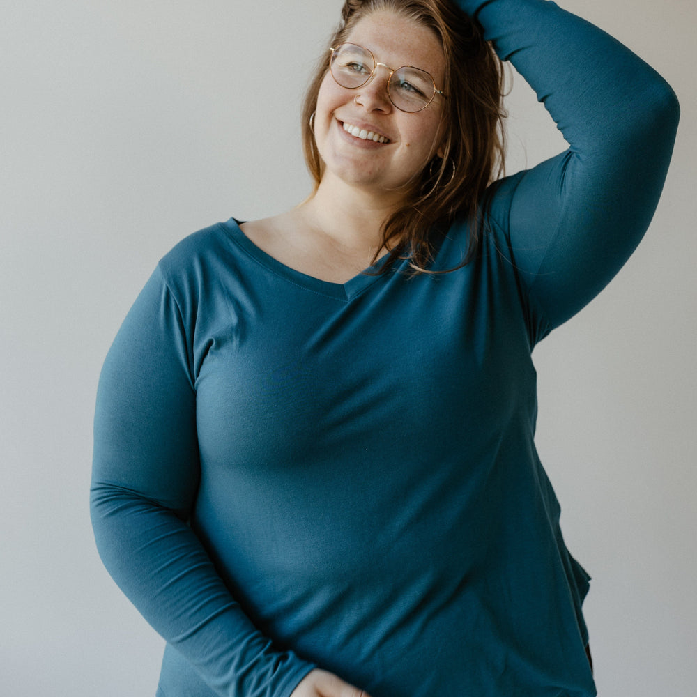 A person in glasses and a slim-fit teal long-sleeve shirt stands against a plain background, smiling with one hand on their head.