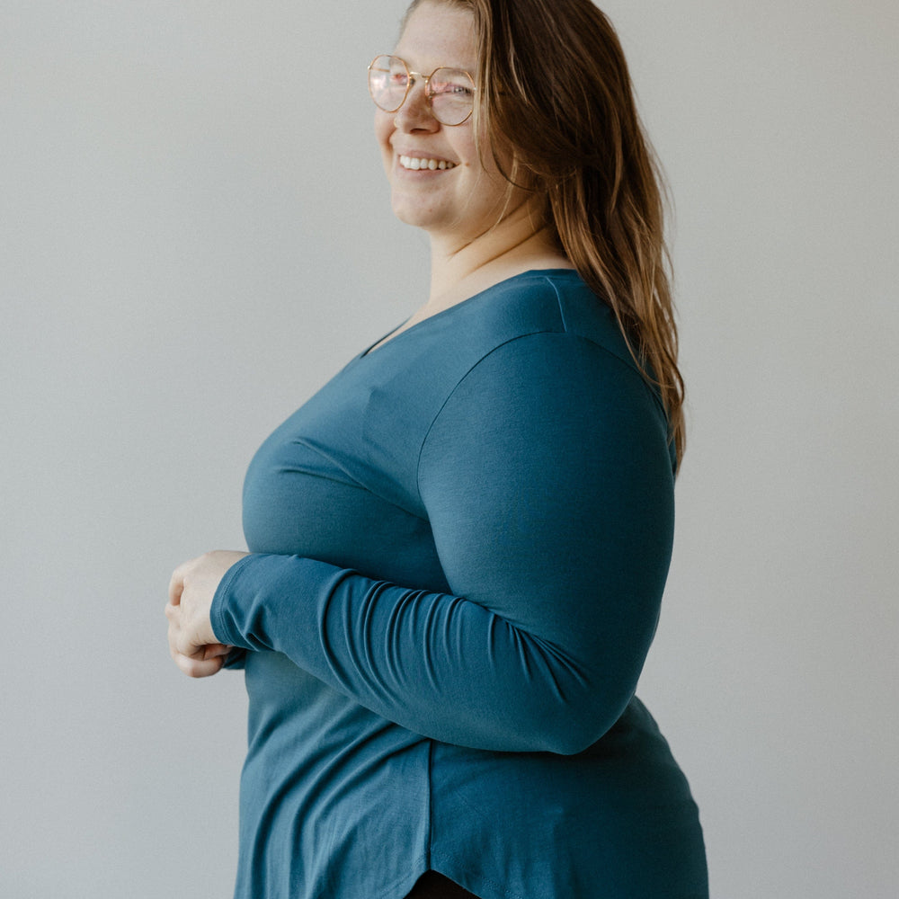 A person wearing glasses and a slim fit teal long-sleeve top is smiling and standing against a plain background.