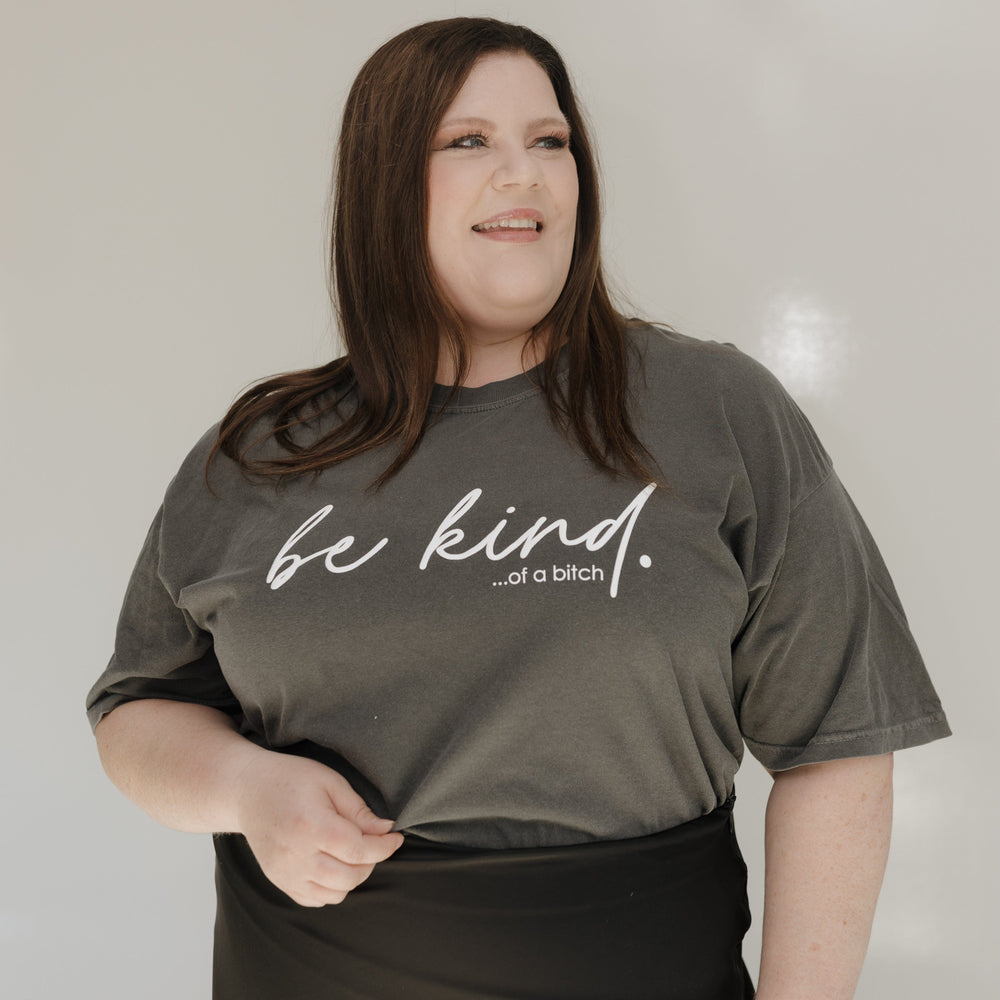 Person wearing a gray t-shirt with 'be kind...of a bitch' text against a white wall.