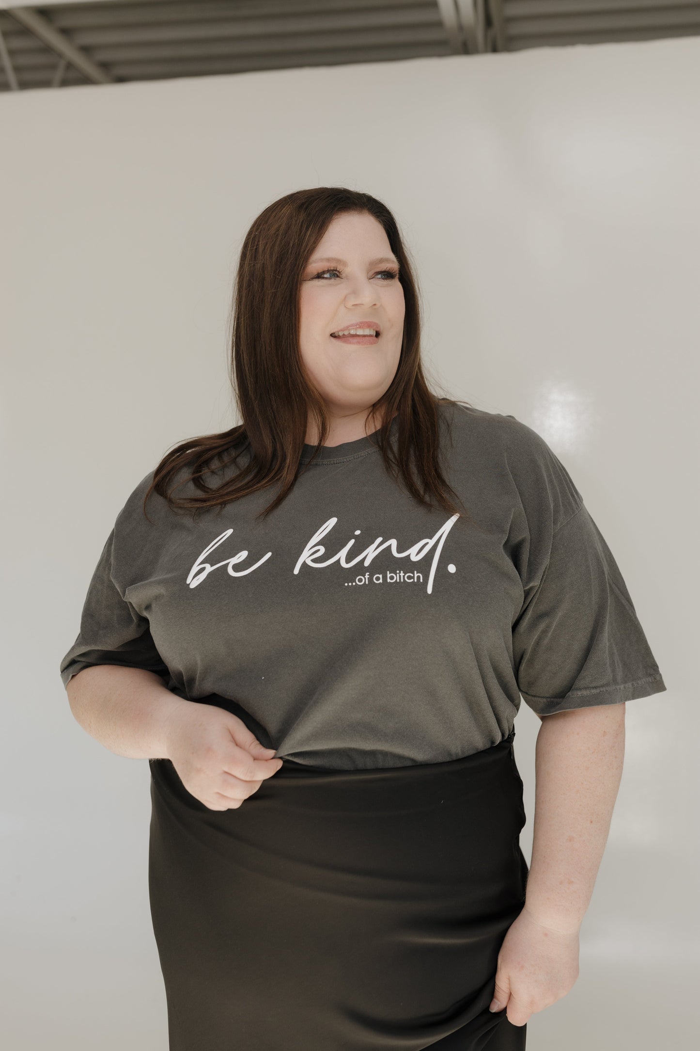 Person wearing a gray t-shirt with 'be kind...of a bitch' text against a white wall.