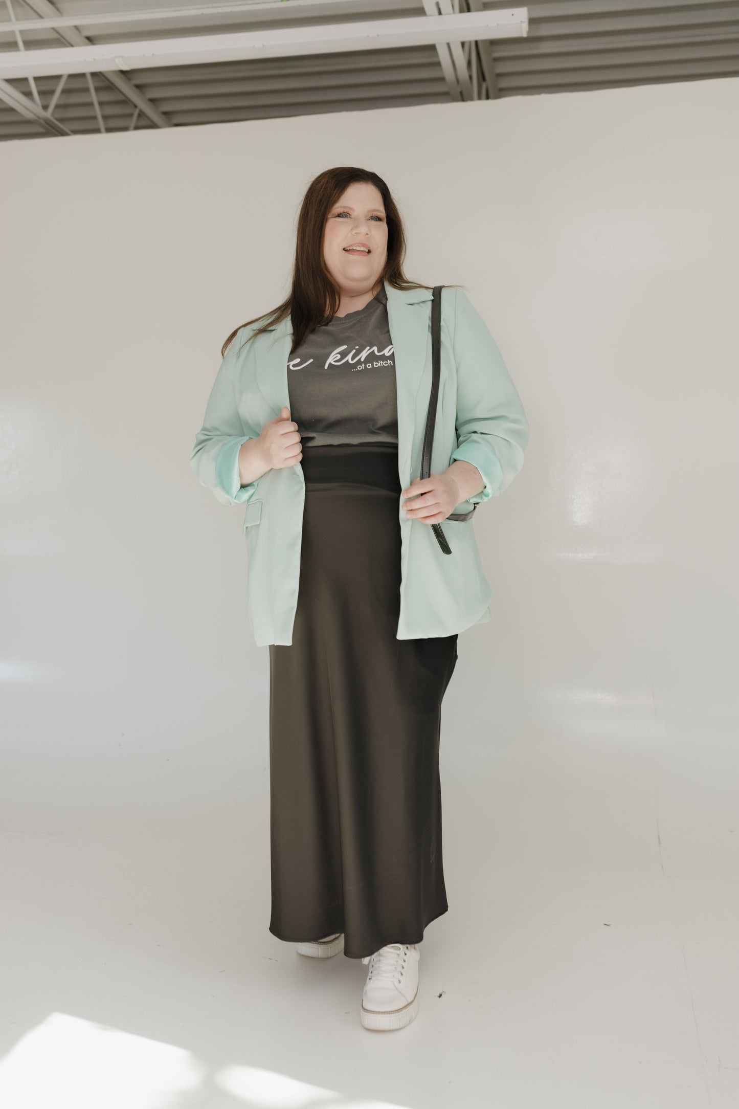 Person wearing a light green jacket, black shirt, and black satin skirt in a white room.
