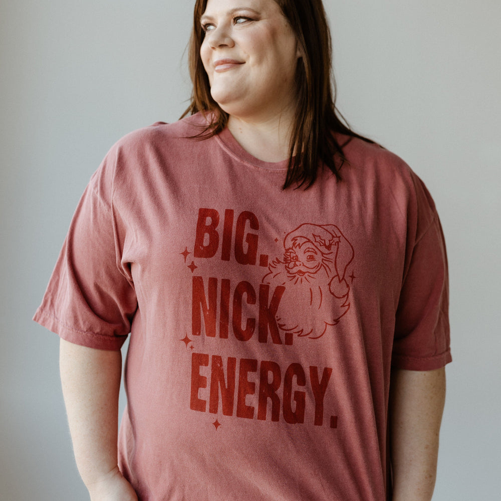 A woman wearing a red graphic tee with "BIG NICK ENERGY" printed on it stands smiling against a plain background. This garment-dyed shirt adds a trendy flair to her vibrant look.
