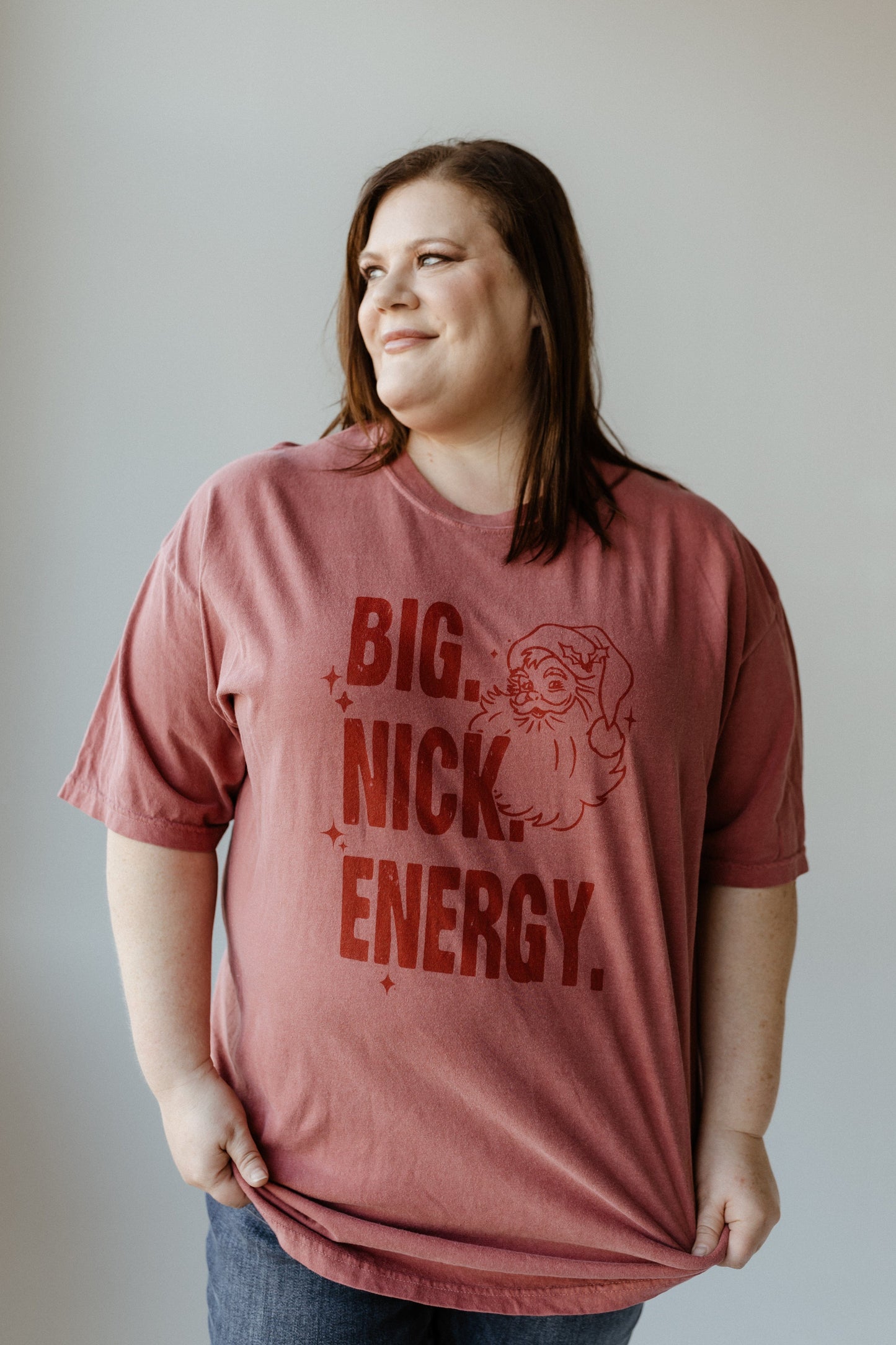 A woman wearing a red graphic tee with "BIG NICK ENERGY" printed on it stands smiling against a plain background. This garment-dyed shirt adds a trendy flair to her vibrant look.