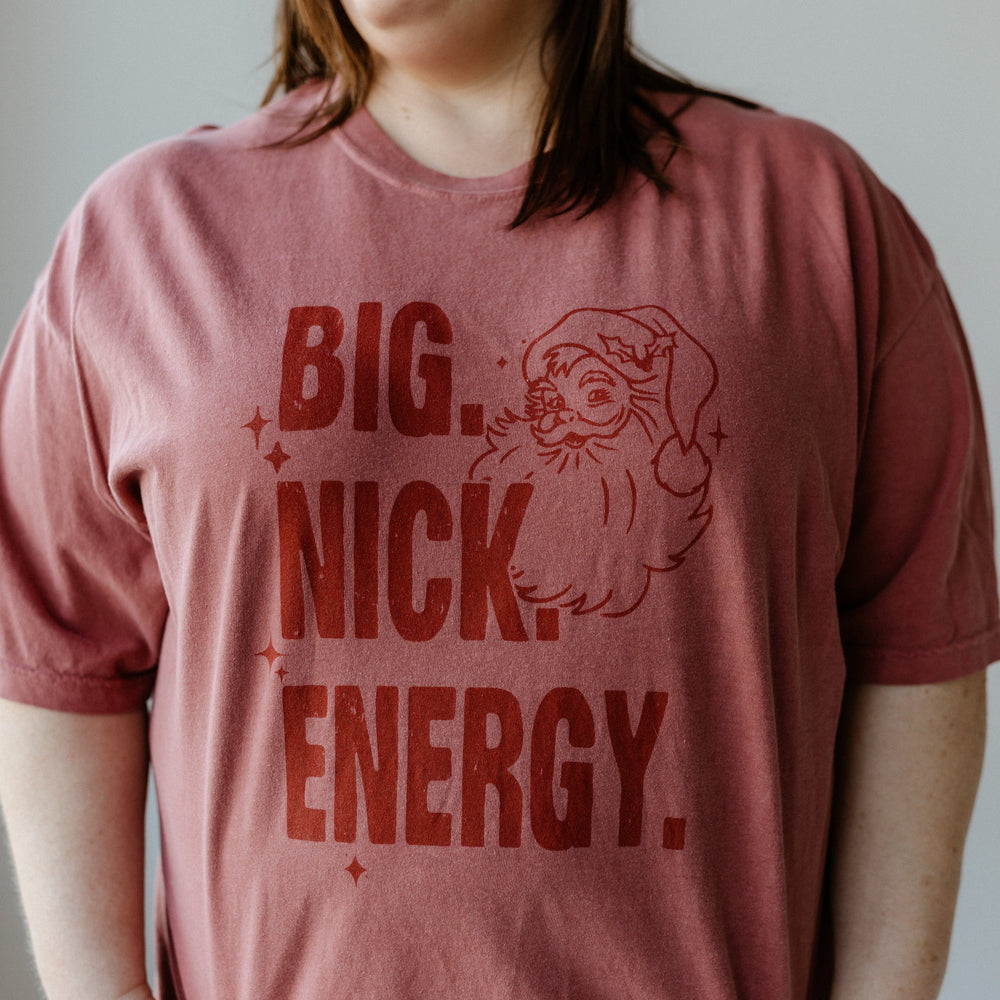 A person wearing a garment-dyed shirt featuring a Santa graphic with the bold text "BIG. NICK. ENERGY." This festive graphic tee adds a playful twist to holiday fashion, perfect for making a jolly statement.