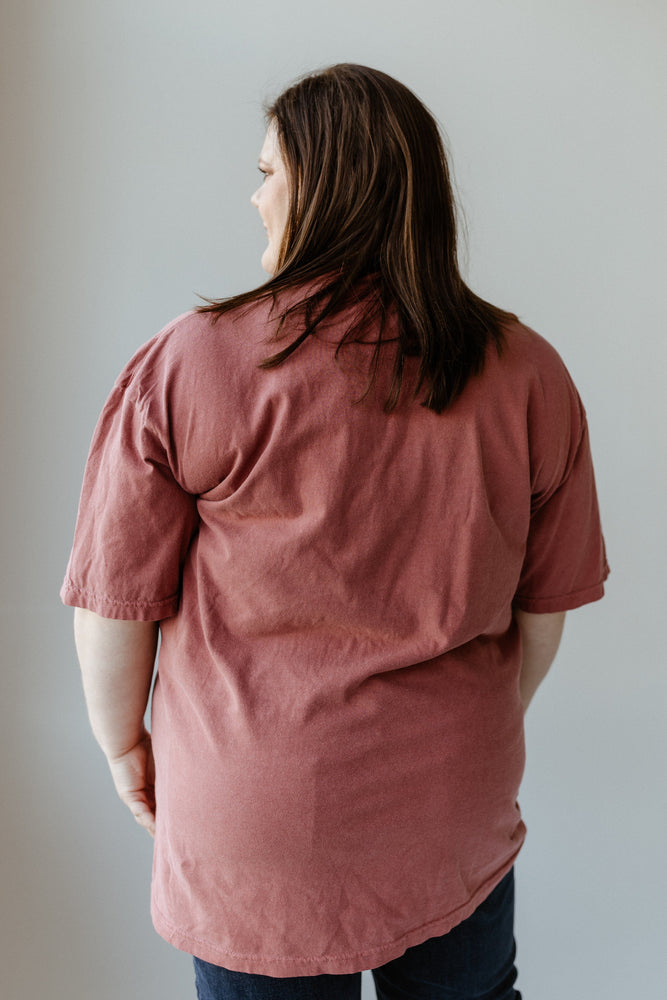 A person with long hair stands with their back to the camera, showcasing a maroon graphic tee paired with dark pants against a plain background.