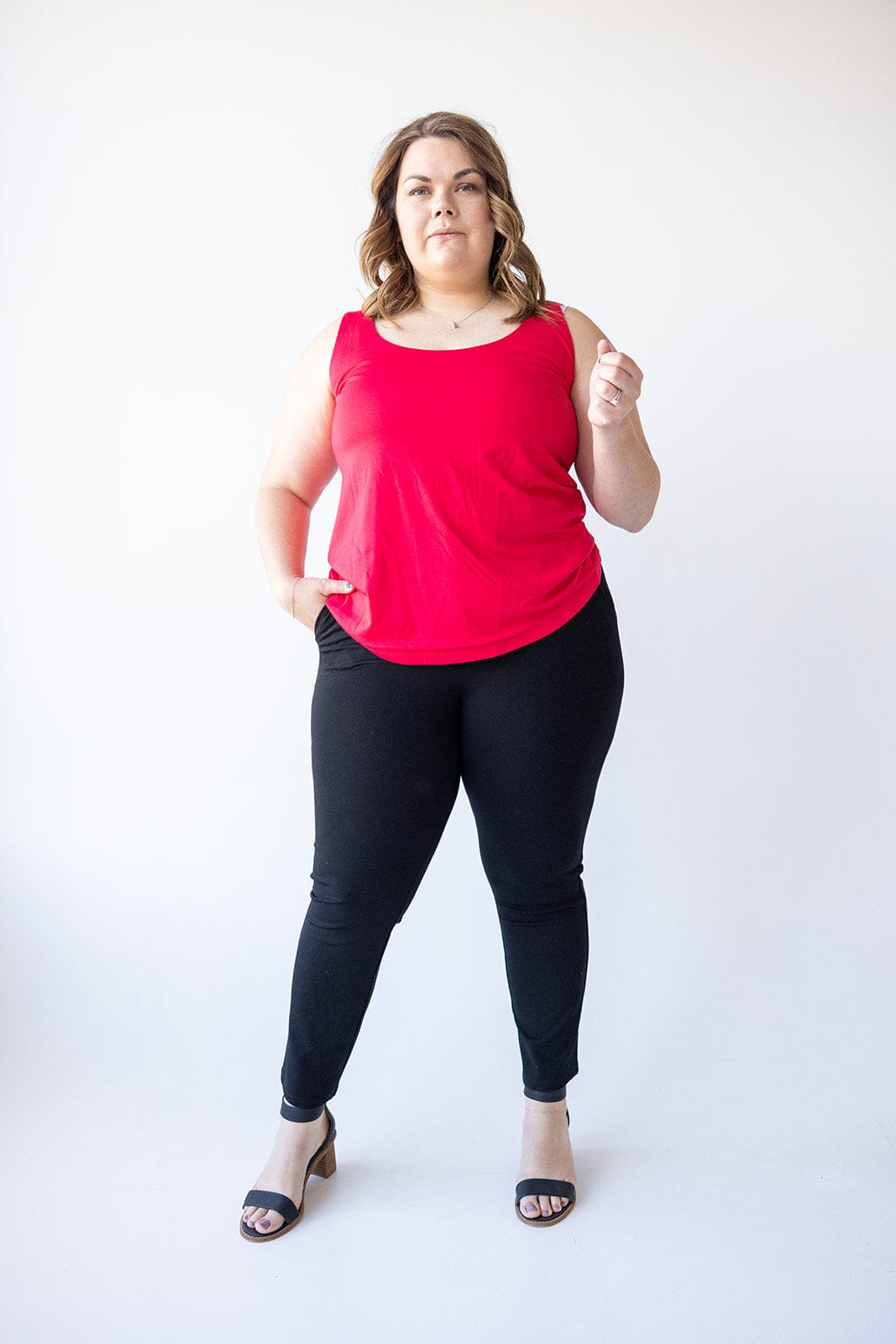 A woman in a red sleeveless top and elegant black dress pants, made from a luxury fabric, stands against a white background.