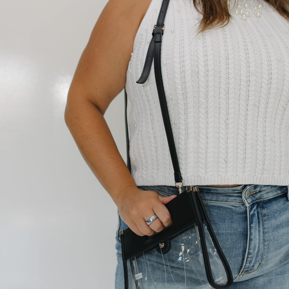 Person wearing a white sleeveless top and denim shorts, holding a black crossbody bag against a plain background.