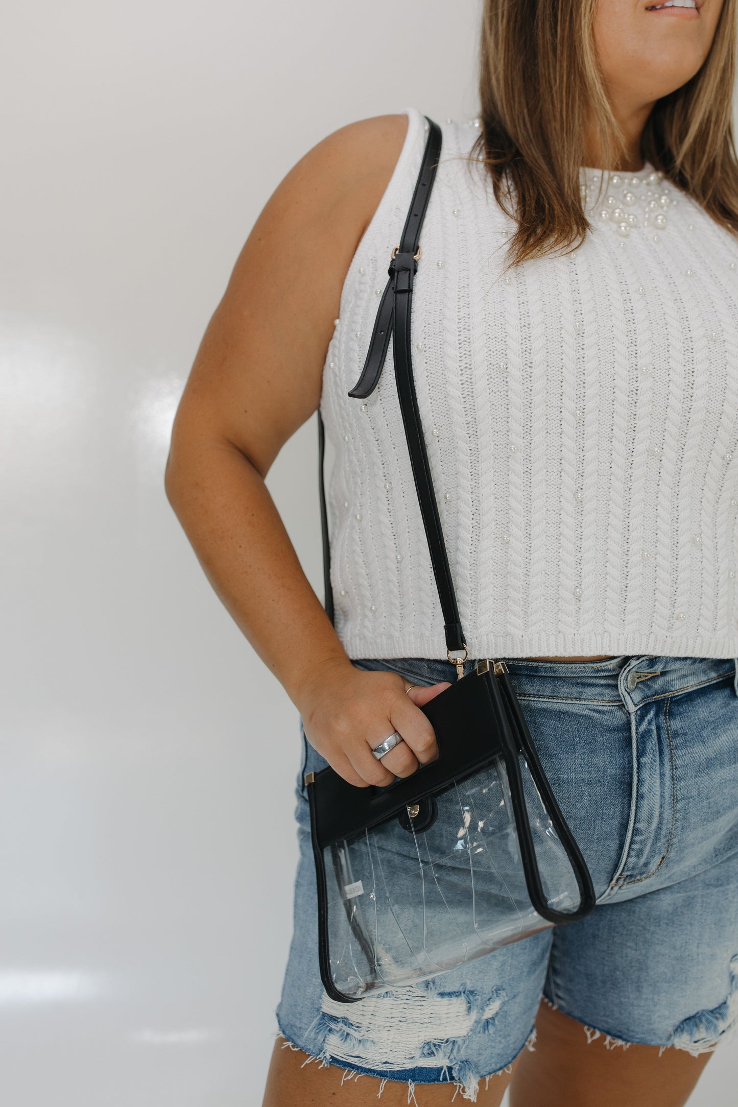 Person wearing a white sleeveless top and denim shorts, holding a black crossbody bag against a plain background.