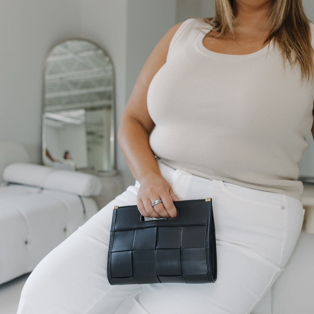 Woman holding a black woven clutch in a minimalistic room.