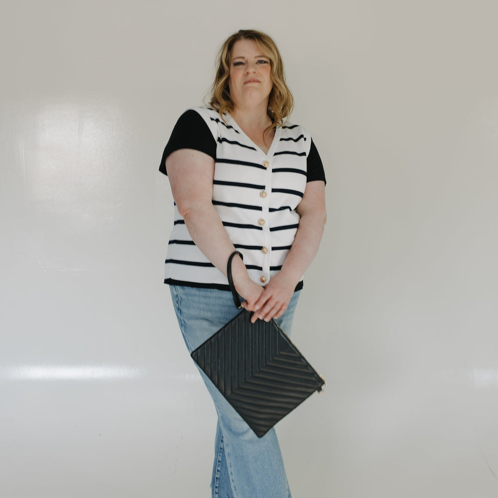 Woman wearing a black and white striped shirt, blue jeans, and white sneakers holding a black clutch against a white background.