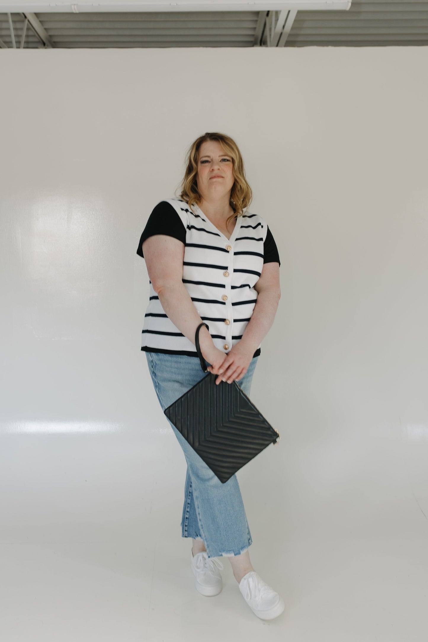 Woman wearing a black and white striped shirt, blue jeans, and white sneakers holding a black clutch against a white background.