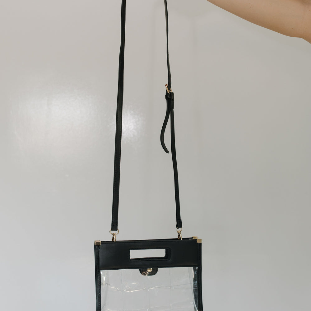 Clear handbag with black strap held by a hand against a light gray background