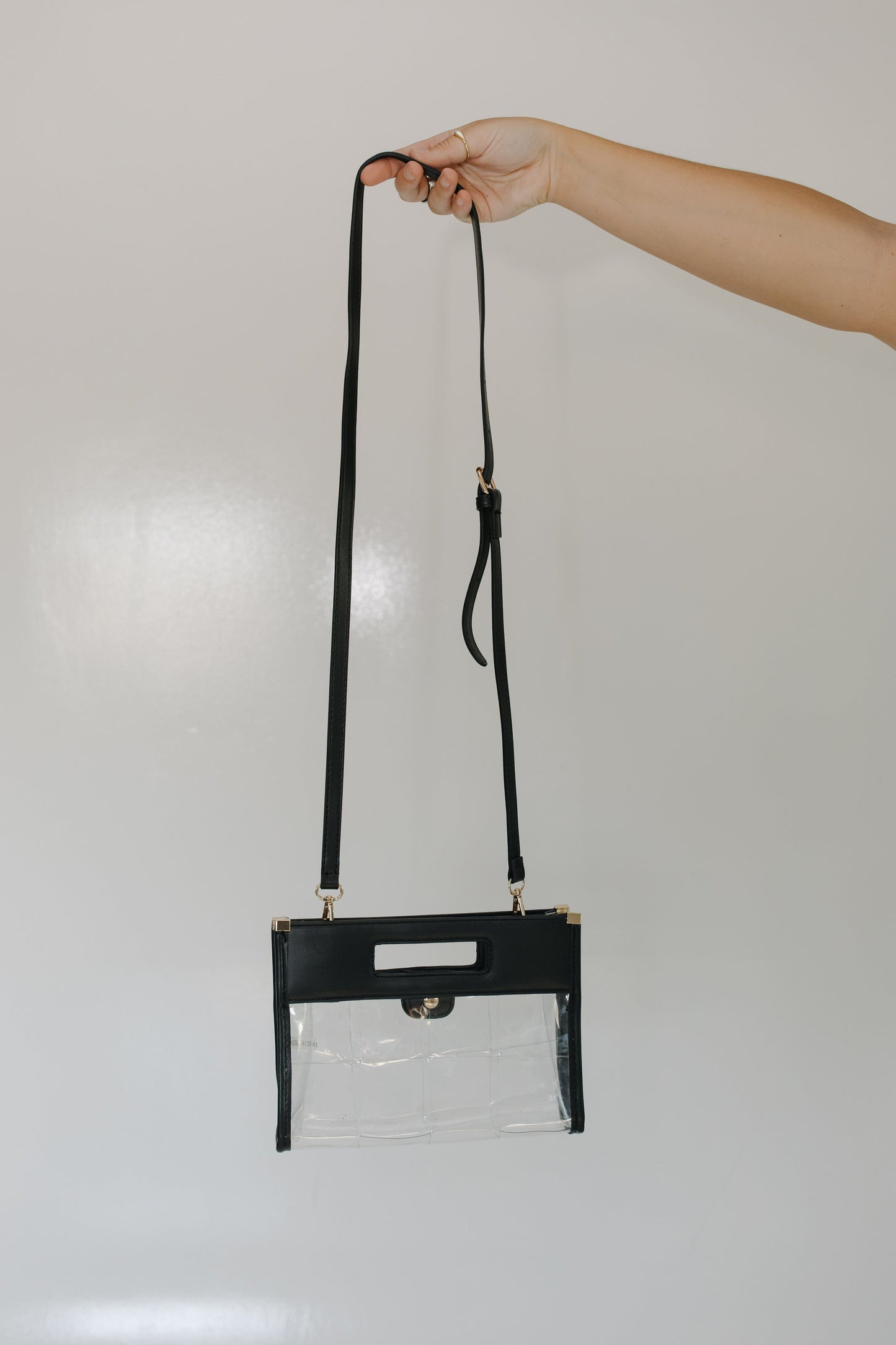 Clear handbag with black strap held by a hand against a light gray background