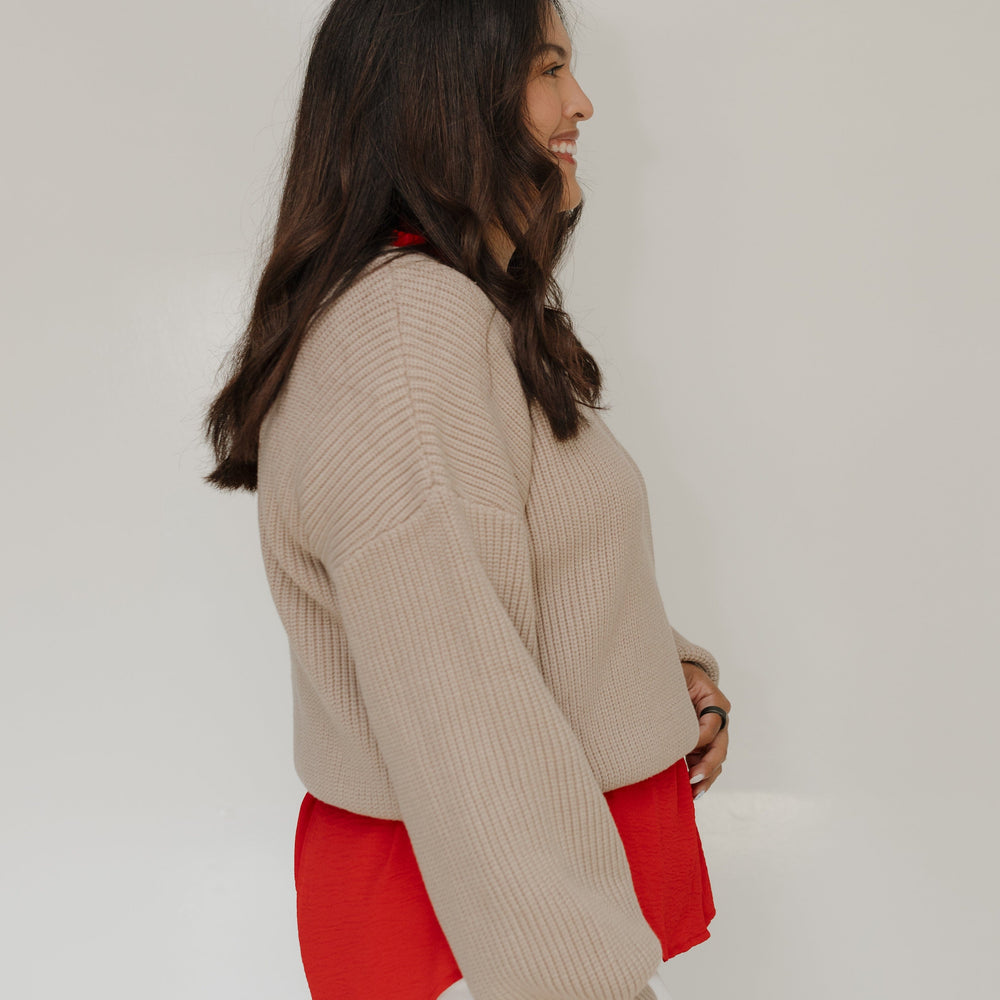 Woman wearing a beige sweater over a red shirt with white pants on a plain background