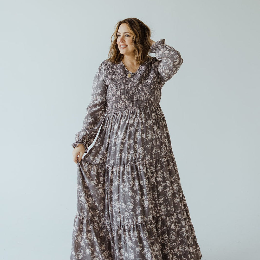 A woman stands against a plain background, wearing an elegant BOHO FLOOR LENGTH FLORAL DRESS IN OPAL GREY by Dex. She has one hand up near her hair and the other lightly holding her dress. She is smiling slightly.