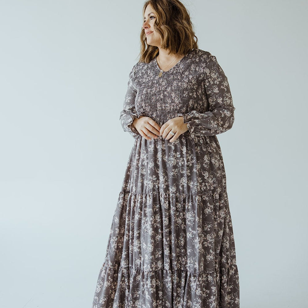 A woman wearing the Dex BOHO FLOOR LENGTH FLORAL DRESS IN OPAL GREY stands against a plain background, looking off to the side.