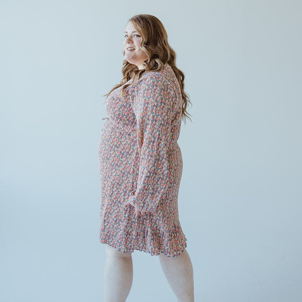 A person with long hair stands sideways, smiling, wearing a BOHO FLORAL DRESS WITH RUFFLE HEM IN FESTIVE BLOOM by Love Marlow and black flats against a plain background.